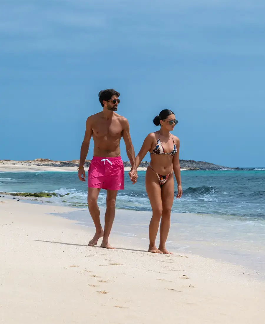 A couple holding hands and walking on the beach wearing swimsuits